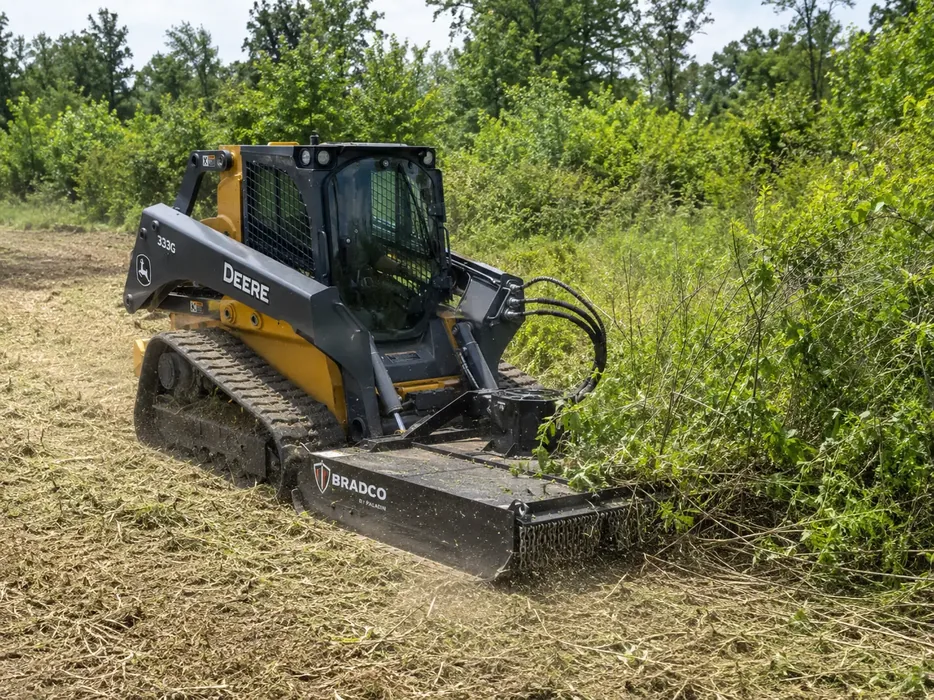 Rotary brush cutter on a compact track loader cutting tall brush in a field, brush hogging
