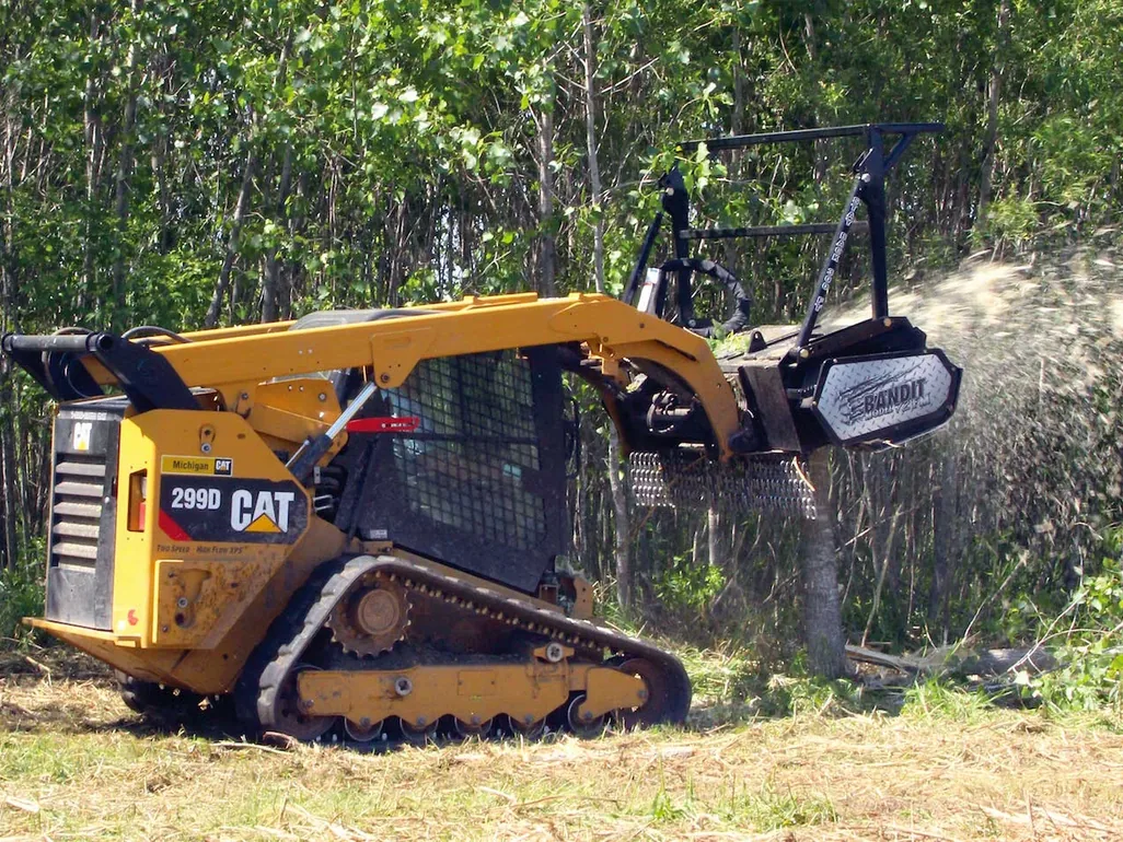 Compact track loader with a mulching head grinding saplings at a woods edge